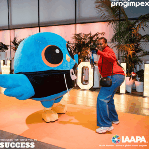 Person in a red jacket posing with a large blue cartoon bird mascot at an indoor event, with plants and illuminated letters in the background.